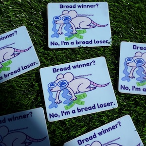 May include: A rectangular white sticker featuring a cartoon illustration of a crying light purple mouse lying on green dollar bills. The text reads Bread winner? No, I'm a bread loser. and includes the social media handle @thelittleriverrat.