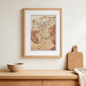 Puede incluir: Impresión artística enmarcada de un mapa vintage con marco de madera clara. El mapa presenta una ilustración detallada en tonos marrones, rojos y beige, que representa un paisaje con edificios y montañas. La obra de arte se exhibe sobre una superficie de madera.