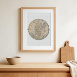 May include: A framed, circular map print with a light wood frame. The map features a detailed, vintage design with various colours. A small, beige bowl and a wooden chopping board are on the wooden surface below the framed print.