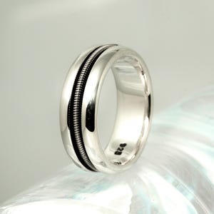 May include: A silver ring with a central black band, possibly a guitar string design. The ring has a polished, reflective surface and a simple, elegant style. The ring is a jewelry item.