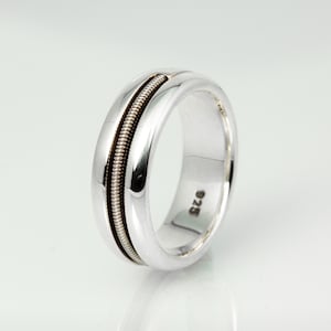 May include: A silver ring with a guitar string inlay. The ring has a polished, rounded band with a groove that holds the string. The ring is marked with "925" on the inside, indicating sterling silver. A unique piece of jewelry.