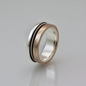 May include: A silver and rose gold wedding band with a textured finish. The band is made of two metals, with the rose gold band being on top of the silver band.