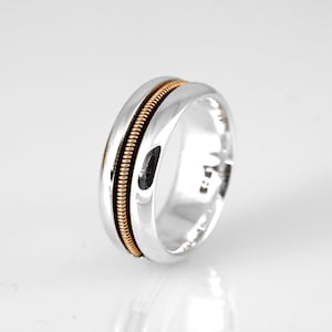 May include: A silver ring with a gold-colored, textured band running through the center. The ring has a polished, reflective surface, and the gold band adds a contrasting detail to the design.