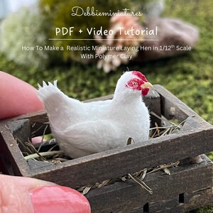 Op de afbeelding: Een miniatuur witte kip met rode accenten zit in een kleine houten kist gevuld met stro. De kip maakt deel uit van een tutorial over het maken van een realistische miniatuur legkip op schaal 1/12 met polymeerklei. De tekst "Debbieminiatures PDF + Video Tutorial" is zichtbaar.