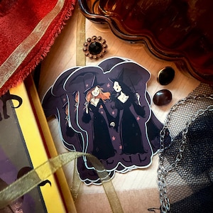 May include: A die-cut sticker featuring an illustration of two witches in black dresses holding hands under a dark purple sky with stars. The witches wear pointed hats and carry umbrellas. The sticker is surrounded by red ribbon, a gold button, and a chain.