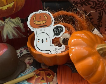 May include: A white ghost holding a jack-o'-lantern with the word "BOO!" in a black speech bubble. The ghost and pumpkin are on a sticker with an orange pumpkin background.