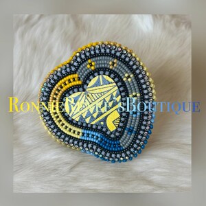 May include: Heart-shaped beaded item with a yellow, blue, and gray color scheme. The center features a yellow design on a wooden base. The item is surrounded by a beaded border. The text "RONNIE'S BOUTIQUE" is visible.