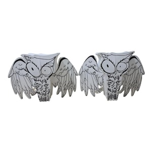 May include: Two white owl figurines featuring black line art illustrations of feathers and large circular eyes. Each owl is depicted with wings spread wide to the sides, showcasing detailed ink-style drawings on a smooth, solid white surface.