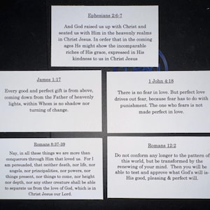 May include: Five white rectangular cards with black text, each displaying a Bible verse. Verses include Ephesians 2:6-7, James 1:17, 1 John 4:18, Romans 8:37-39, and Romans 12:2. The cards are arranged on a dark surface.