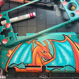 May include: A teal handheld gaming console shell featuring a hand-painted orange Charizard dragon with blue wings and flames. The shell includes a matching teal faceplate with red circular icons and orange paint splatters throughout the design.