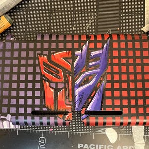 Könnte beinhalten: Maßgefertigte Nintendo Switch Joy-Con-Controller mit den Autobot- und Decepticon-Symbolen aus Transformers. Die Controller sind rot, lila und schwarz mit einem Rastermuster.