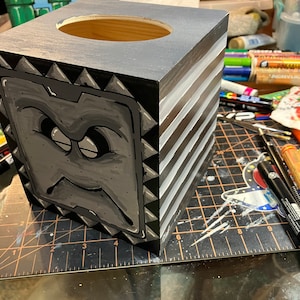 Op de afbeelding: Een zwarte houten tissuebox met een wit en zwart gestreept ontwerp. De voorkant van de doos heeft een cartoon gezicht met een fronsende uitdrukking. De doos heeft een vierkante opening aan de bovenkant voor tissues.