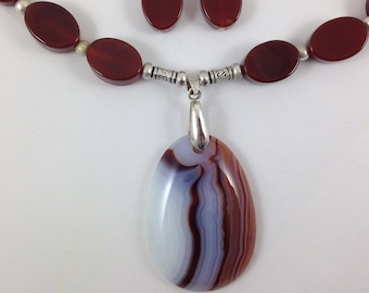 May include: A red agate necklace with a silver pendant featuring a white and brown agate stone. The necklace has silver beads and matching earrings.