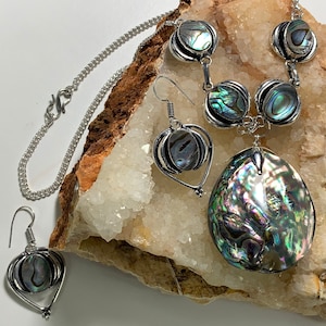 Könnte beinhalten: Ein silbernes Halsketten- und Ohrring-Set mit schillernden Abalone-Muschel-Anhängern. Die Halskette hat einen Karabinerverschluss und die Ohrringe haben Hakenverschlüsse.