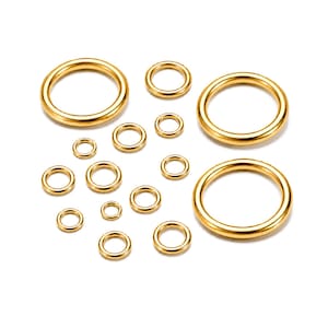 Puede incluir: Una colección de catorce anillos metálicos circulares de tono dorado, lisos y de diámetros variables, dispuestos en un patrón disperso. Cada anillo presenta una superficie pulida y reflectante con una sección transversal redondeada y uniforme.