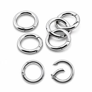 May include: A collection of seven silver-toned metal spring gate rings. These circular hardware components feature a polished, reflective finish and a hinged mechanism that allows the ring to open and close securely for attaching various items.
