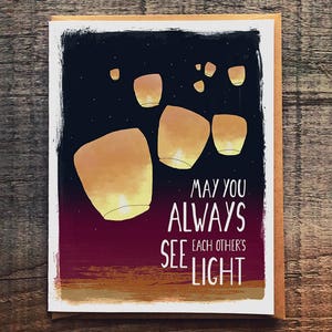 May include: A greeting card with a dark blue background and a design of glowing lanterns in the night sky. The text on the card reads "May you always see each other's light."