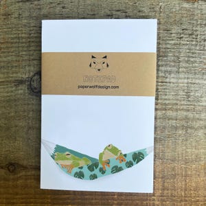 May include: White notepad with a brown band featuring the text "NOTEPAD" and "paperwolfdesign.com". The notepad has a whimsical illustration of two green frogs relaxing in a blue hammock with green leaves.