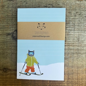 May include: A light blue notepad with horizontal lines, featuring a cartoon cat skiing on a snowy slope. The cat wears goggles, a yellow jacket, and orange pants. The notepad has a brown band with the word "NOTEPAD" and the website "paperwolfdesign.com".