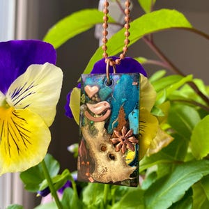 May include: A copper chain necklace with a rectangular pendant featuring a mixed media design. The pendant is made of resin and includes copper, turquoise, and gold accents. The design includes a bird, a heart, and a flower.