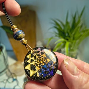 May include: A circular pendant featuring a gold and blue geometric Sri Yantra pattern set in a black resin base with gold flecks. The pendant hangs from a black cord accented by a matching spherical bead and metallic gold-toned hardware components.