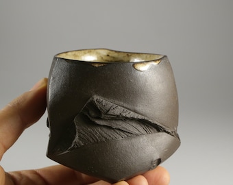 May include: A dark brown ceramic cup with a textured, uneven surface. The cup has a slightly rounded shape and a wide opening.