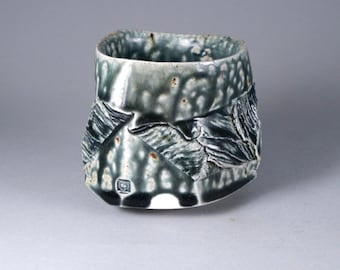 Puede incluir: Una taza pequeña de cerámica blanca con un esmalte texturizado negro y gris. La taza tiene una forma angular única y una pequeña marca negra en la parte inferior.