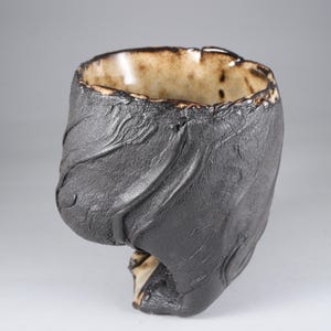 Op de afbeelding: Een handgemaakte keramische beker met een unieke, getextureerde donkergrijze buitenkant. De binnenkant is lichtbruin met een glanzende afwerking. Het ontwerp van de beker heeft organische, vloeiende lijnen, wat een sculpturale esthetiek creëert. De rand heeft een onregelmatige vorm.