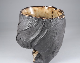 May include: A handmade ceramic cup with a distinctive, textured dark grey exterior. The interior is light brown with a glossy finish. The cup's design incorporates organic, flowing lines, creating a sculptural aesthetic. The rim has an irregular shape.