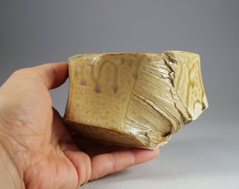 May include: A handcrafted ceramic bowl with a distinctive, textured pattern. The bowl has a light brown hue with darker brown detailing and a rough, irregular surface. The bowl is held in a hand, highlighting its form and size.