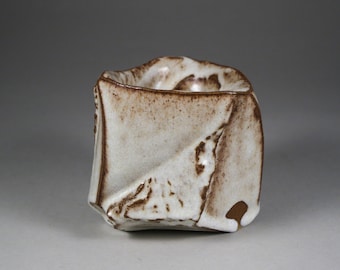 May include: A small, white ceramic cup with a brown glaze. The cup has a unique, folded design and a rough, textured surface.