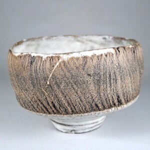 May include: A small, white ceramic bowl with a rough, textured exterior. The bowl has a wide, rounded rim and a slightly flared base.