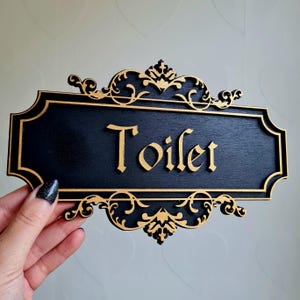 May include: Black wooden sign with gold ornate trim and the word "Toilet" written in gold script.