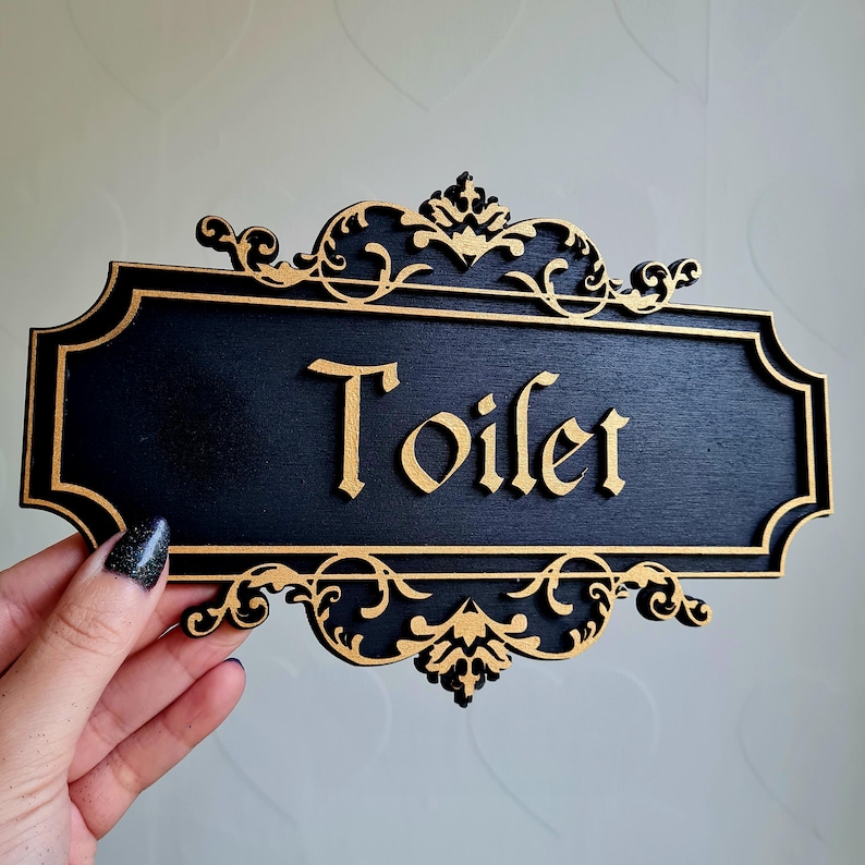 May include: Black wooden sign with gold ornate trim and the word "Toilet" written in gold script.