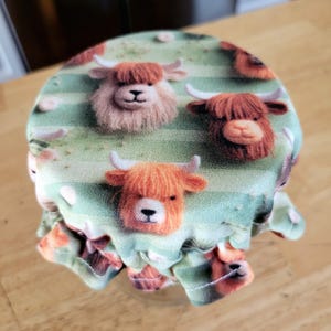 May include: A green and white fabric jar lid with a repeating pattern of brown and white Highland cows. The lid has a ruffled edge.