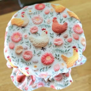 May include: A fabric jar cover with a repeating pattern of chickens and flowers. The chickens are in shades of yellow, orange, and brown, while the flowers are pink and peach. The background is a light, off-white color.