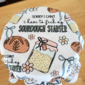 May include: A white fabric jar cover with a floral and bread design. The cover has the text "SORRY I CAN'T I have to feed my SOURDOUGH STARTER". The cover is designed to fit over a jar.