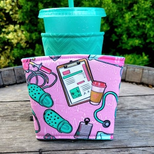 May include: Pink and green reusable cup sleeve with a medical theme. The sleeve features a stethoscope, a clipboard with a medical chart, a pair of green clogs, and a coffee cup.
