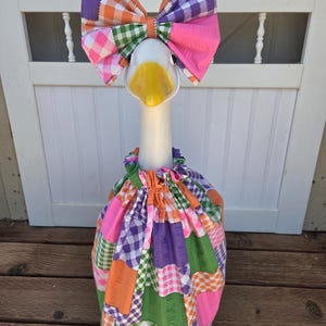 May include: A decorative goose figurine wearing a matching outfit consisting of a large, multi-colored gingham bow headpiece and a gathered, sleeveless dress. The fabric features a patchwork pattern of orange, purple, green, and pink checks.