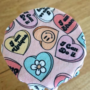 May include: A pink fabric jar cover with a pattern of colorful conversation heart candies. Each heart has a different message, including "I am Strong" and "I Can Do It". A light blue heart has a white flower.