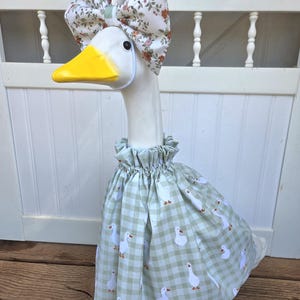 May include: A white decorative goose figurine wearing a light green and white gingham dress featuring a repeating pattern of small white geese. The goose also wears a matching oversized fabric bow on its head with a floral print in orange and green.