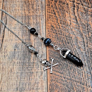 May include: A silver chain with black beads and a black crystal pendulum. The pendulum is attached to a silver skull and crossbones charm.