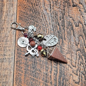 May include: A silver-toned keychain featuring a lobster clasp, a triangular brown glittery stone pendant, and various charms including an artist palette, a bow and arrow, an eye symbol, and small red and metallic beads attached by silver chains.