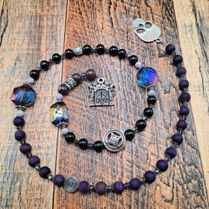 May include: A handcrafted rosary with dark purple and black beads, interspersed with iridescent crystals and silver accents. Features a cemetery gate charm, a skull pendant, and a decorative silver clasp. The rosary is displayed on a wooden surface.