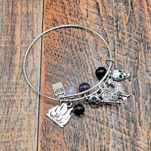 May include: Silver bangle bracelet with various charms, including a skull, a tombstone with "RIP" text, a flame, and black and purple beads. The bracelet is displayed on a wooden surface.