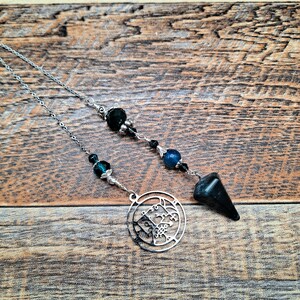 May include: A silver pendulum with a black and blue crystal design. The pendulum has a circular charm with a detailed design and a pointed black crystal at the end.