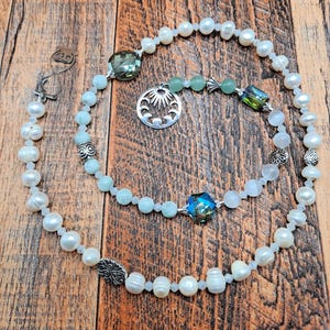 May include: Two layered necklaces with pearls, blue beads, and decorative silver charms. One necklace features a large, faceted green crystal and a sunburst pendant. The other necklace has a large, faceted blue crystal. The necklaces are on a wooden surface.
