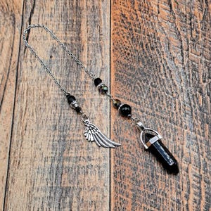 May include: A silver-toned metal chain pendulum featuring a faceted black stone point pendant. The chain is adorned with small black beads and a detailed silver-toned metal wing charm, creating a balanced design with dark and metallic elements.