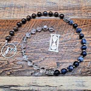 May include: A beaded mala necklace with clear quartz, brown jasper, and blue and black jasper beads. The necklace has a silver charm with an hourglass design and a silver charm with a mountain design.