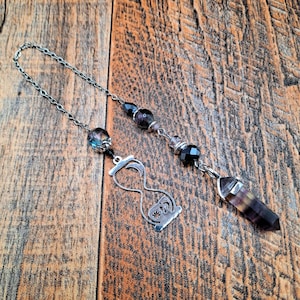 May include: A silver-toned metal chain pendulum featuring a faceted purple and green crystal point, a silver hourglass charm with moon and star cutouts, and several small black and iridescent glass beads arranged along the chain links.